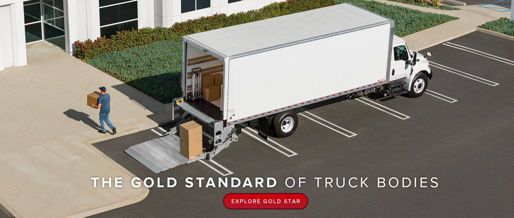 A large, white dry freight truck is parked in front of a corporate building. A delivery man is carrying a box to the front door.