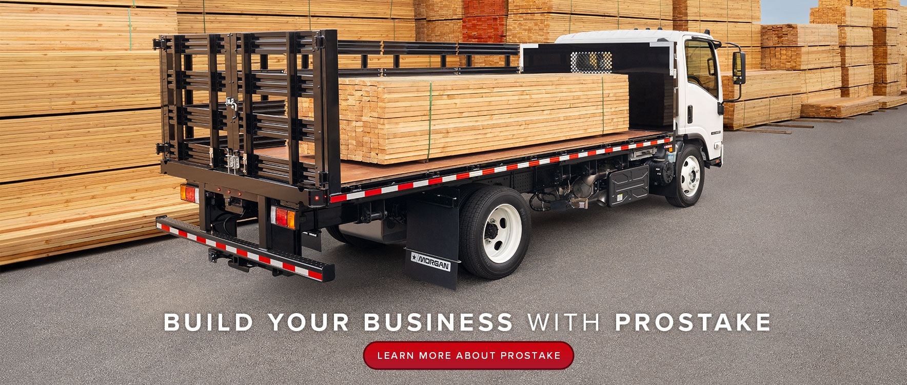 A white flatbed truck with black stakes is parked, loaded with wood, surrounded by stacks of wood in a large lumberyard.
