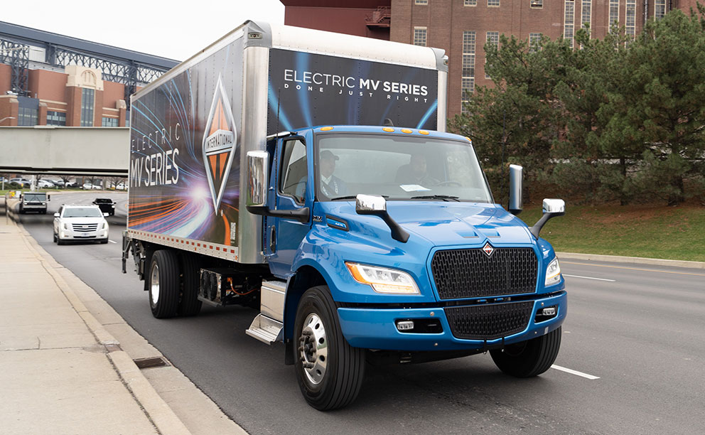 Large dry freight body with full-body decals, mounted on a blue International MV Series chassis, photographed from the front, while driving on a highway.